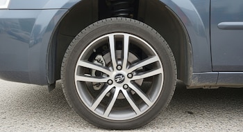 Rueda de automóvil con llanta de radios grises y neumático negro, freno de disco, pinza de freno y amortiguador visible bajo un guardabarros gris oscuro sobre asfalto.