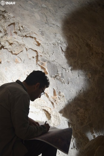 Hombre de perfil con gafas escribe en un mapa o plano. Pared rocosa clara, parcialmente iluminada y con sombras. Logotipo INAH