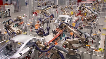 Vista aérea de una planta de fabricación con múltiples robots metálicos de brazos articulados ensamblando carrocerías de vehículos. Un operario humano está en el fondo.