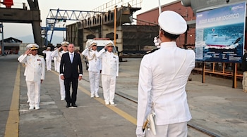 Grupo de militares con uniforme naval blanco y un hombre de traje oscuro en explanada industrial con grúas y cartel de buque logístico