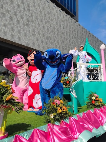 Una mujer con corona y banda blanca saluda desde una carroza decorada con césped artificial y flores, junto a Stitch, Ángel y Lilo disfrazados