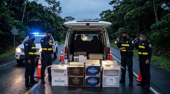 Policías revisan cajas de cigarrillos en baúl de camioneta blanca en carretera de Costa Rica. Conos y luces de patrulla. Fondo de vegetación.