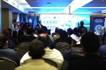Salón con audiencia de espaldas, hombre hablando en un podio, pantalla grande con gráficos y texto, proyector en el techo y varias banderas
