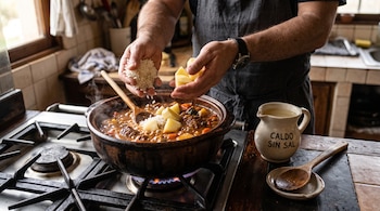 Manos de una persona agregan arroz y papas a una olla con guiso sobre el fuego, al lado de una jarra de caldo y una cuchara.