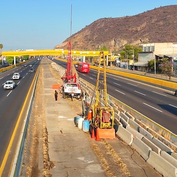 Equipo de perforación amarillo y rojo con un camión blanco y un trabajador en el camellón central de la autopista México-Puebla, con vehículos y una montaña al fondo