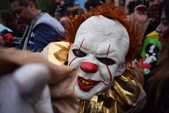 Primer plano de una persona con máscara blanca de payaso, cabello naranja y cuello dorado entre una multitud al aire libre