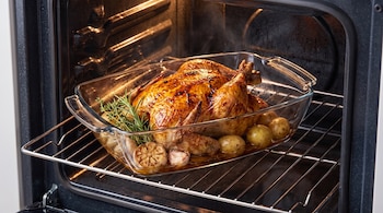 Pollo dorado en recipiente de vidrio sobre la rejilla de un horno, junto a patatas pequeñas, una cabeza de ajo y ramas de romero.