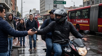 Un hombre en motocicleta arrebata un teléfono a un peatón en una calle lluviosa de Bogotá. Personas con expresiones de sorpresa y un bus TransMilenio son visibles.