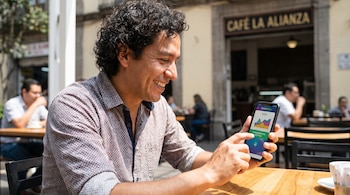 Hombre latinoamericano sonriente con camisa estampada mira su smartphone. La pantalla muestra "Transferencia Exitosa" en una app de banca móvil. Sentado en mesa de madera.