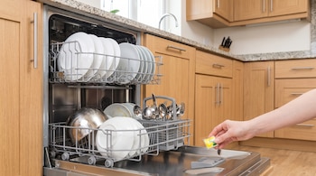 Lavavajillas abierto con dos cestas, platos, cuencos, cubiertos. Una mano sostiene una pastilla de detergente. Fondo de cocina con muebles de madera.