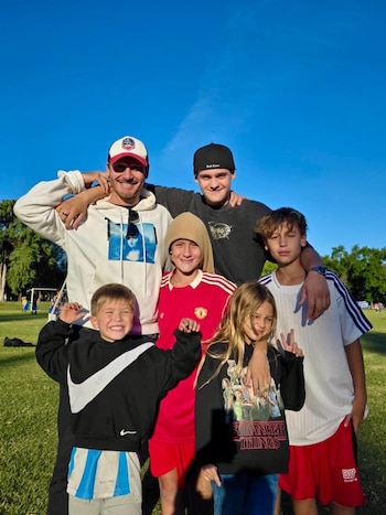 Benjamín Vicuña posa sonriente con sus cinco hijos, todos de pie en un campo de hierba bajo un cielo azul despejado, vestidos con ropa informal y deportiva