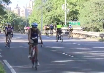 Primer plano de ciclista en bicicleta de ruta, seguido por otros ciclistas en carretera asfaltada con árboles, edificios y una señal de calle al fondo