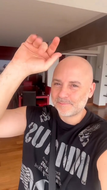 Hombre calvo con barba sonríe a cámara, vistiendo una camiseta negra con diseño gris. Tiene la mano derecha levantada, con índice y meñique extendidos