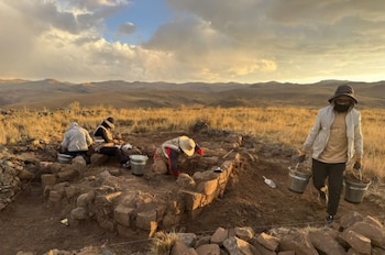 Arqueólogos trabajan en una excavación con estructuras de piedra en un paisaje montañoso mientras uno traslada baldes con tierra