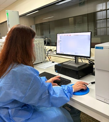 Una científica con bata azul y cabello castaño trabaja en un laboratorio, usando un ordenador y equipo analítico blanco para la detección de sustancias