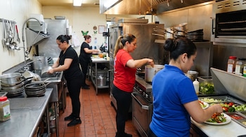 Cuatro mujeres trabajan en una cocina industrial con encimeras de acero inoxidable, fogones con ollas y platos con ensaladas.
