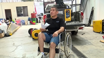 Un hombre sentado en una silla de ruedas sostiene hojas de papel frente a una camioneta blanca, rodeado de maquinaria y herramientas.