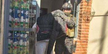Agente policial táctico detiene a una persona con chaqueta sobre la cabeza. Fachada de vivienda con rejas, cortina infantil, columna de ladrillos y maceta colgante