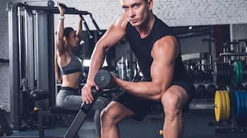 Un hombre de torso musculoso levanta una mancuerna sentado en un banco de gimnasio, con una mujer usando una máquina de poleas en el fondo.