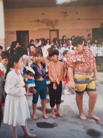 Niños descalzos con disfraces en el patio de una escuela, frente a un grupo de personas y alumnos con guardapolvos blancos