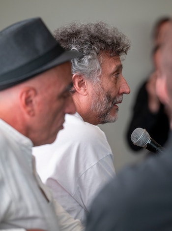 Primer plano de Fito Páez, con cabello canoso y barba, mirando hacia la derecha, junto a un micrófono, y otra persona en primer plano