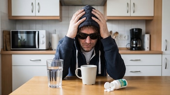 Hombre con sudadera oscura y gafas de sol, con las manos en la cabeza, sentado en una mesa de cocina. Un vaso de agua y un frasco de aspirinas están en la mesa.
