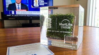 Un cubo de acrílico con césped y tierra, logos de Copa Mundial FIFA 2026 y MetLife Stadium. Un USB FIFA y un certificado de autenticidad sobre una mesa de madera.