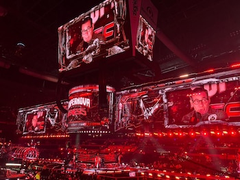 Vista panorámica de un escenario de evento con múltiples pantallas grandes mostrando a un hombre con gafas y micrófono, rodeado de luces rojas y una multitud