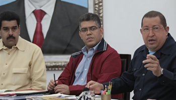 Tres hombres sentados en una mesa, con material de oficina y un micrófono. Al fondo, un retrato de un traje con corbata roja y camisa blanca