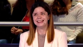 Primer plano de una mujer joven con cabello castaño claro y lacio, sonriendo con un micrófono discreto en su mejilla. Viste un blazer claro