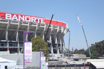 Vista exterior del Estadio Azteca en remodelación con el letrero "BANORTE" visible, grúas de construcción y pancartas en primer plano bajo un cielo azul claro