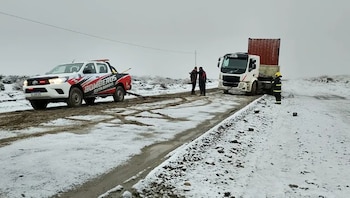 Camioneta blanca de bomberos remolcando un camión blanco con un contenedor rojo en una carretera nevada y embarrada, con personas en la escena