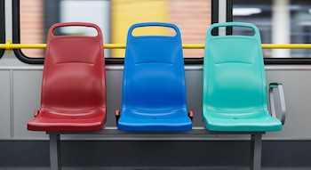 Tres sillas de plástico en un autobús, una roja a la izquierda, una azul en el centro y una aguamarina a la derecha, con ventanas y barra amarilla al fondo.