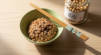 Un bol verde con natto y palillos sobre una mesa de madera clara. Un tarro de vidrio con granos de soja y una etiqueta con kanji japonés está a la derecha.