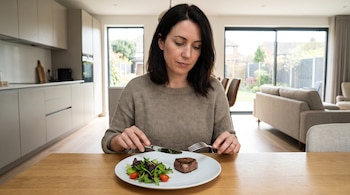 Una mujer sostiene un tenedor y un cuchillo frente a un plato blanco con un trozo de carne y ensalada sobre una mesa de madera.
