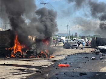 Varios socorristas con uniformes blancos y rojos inspeccionan un automóvil negro destruido junto a la estructura de un camión