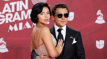 Ángela Aguilar con vestido brillante y Christian Nodal con traje negro y gafas de sol posan frente a un fondo rojo con logotipos.