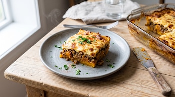 Porción cuadrada de pastel de calabaza y carne en un plato gris junto a una fuente de vidrio y una espátula sobre una mesa de madera.