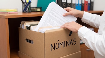 Manos de una persona con camisa blanca colocando folios impresos en una caja de cartón que tiene escrita la palabra NÓMINAS.
