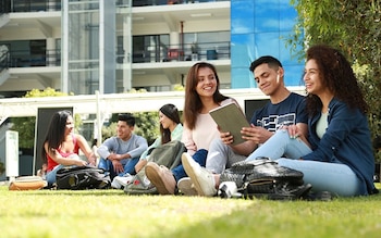 Seis jóvenes, cinco mujeres y un hombre, sentados en el césped de un campus universitario con mochilas y un dispositivo electrónico