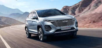 Un SUV plateado, modelo Chery Tiggo 2 Pro, transita una carretera con montañas y cielo azul de fondo. Pequeñas aves vuelan en la distancia