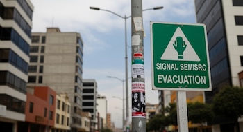 Un cartel verde y blanco de "SIGNAL SEMISRC EVACUATION" en primer plano de una calle urbana peruana con edificios y un poste de luz bajo un cielo nublado.