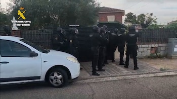 Agentes de la Guardia Civil con uniforme táctico oscuro están ante la entrada de una casa, junto a un vehículo blanco estacionado en la calle