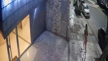 Un edificio con ventanales y pared de piedra. Una acera con una mujer caminando. Una calle con un coche blanco aparcado y un paso de cebra