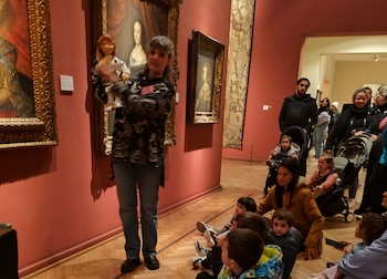 Una mujer con una muñeca interactúa con niños sentados en el suelo y adultos de pie. En el fondo, paredes rojas exhiben cuadros y tapices en el museo