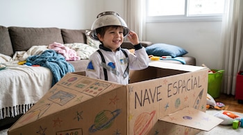 Un niño sonriente con traje de astronauta y un colador en la cabeza está sentado dentro de una caja de cartón decorada como nave espacial.