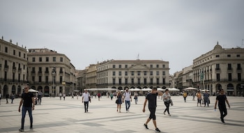 Plaza de ciudad rodeada por edificios clásicos de piedra. Numerosas personas caminan por la plaza bajo un cielo nublado y difuso.