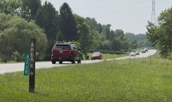 Carretera asfaltada con un SUV rojo y varios coches. Poste de madera con 'AREA K 8260' y señal verde a la izquierda. Hierba, árboles y torre de electricidad