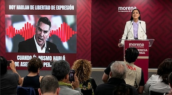Una mujer en un podio con logo de Morena y micrófonos. Detrás, una pantalla muestra a un hombre y texto. Varias personas observan desde el público