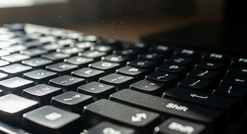 Primer plano de un teclado de computadora negro donde la luz del sol incide sobre las teclas, revelando partículas de polvo suspendidas en el aire.